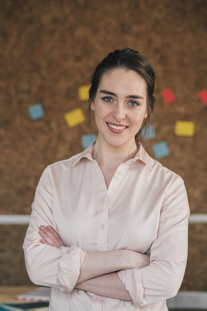 Portrait of a female office worker. She is standing in front of a cork board, smartly dressed with her arms folded and is smiling for the camera.の写真素材