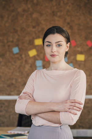 Portrait of a female office worker. She is standing in front of a cork board, smartly dressed with her arms folded and is smiling for the camera.の写真素材