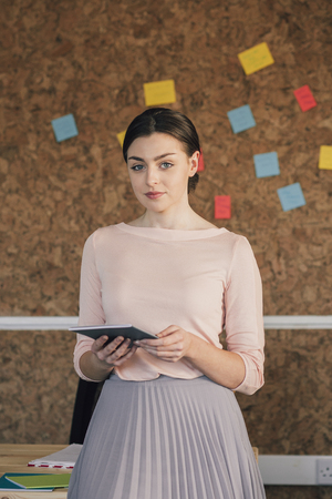 Portrait of a female office worker. She is standing in front of a cork board, smartly dressed with a digital tablet and smiling for the camera.の写真素材