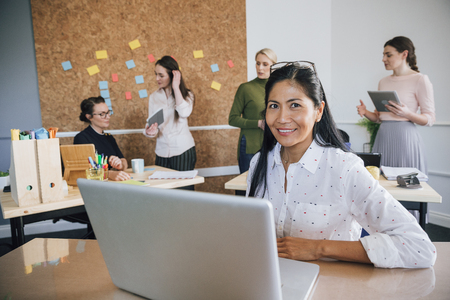 Beautiful woman is smiling for the camera while sitting at her desk in her workplace. There are women working in the background.の写真素材