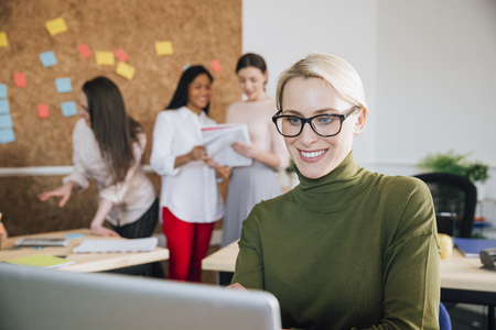 Beautiful woman is smiling while working at her desk in her workplace. There are women working in the background.の写真素材