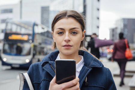 Portrait of a young woman with a smart phone. She is standing in the city and is looking at the camera.の写真素材