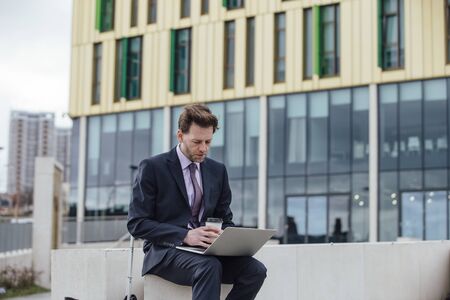 Businessman is working in the city. He is using a laptop and drinking a coffee.