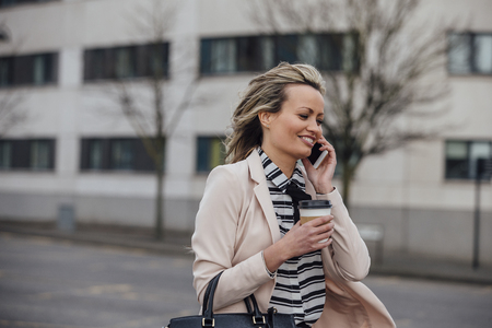 Businesswoman is walking to work in the city. She is holding a coffee cup and is talking on the phone.の写真素材