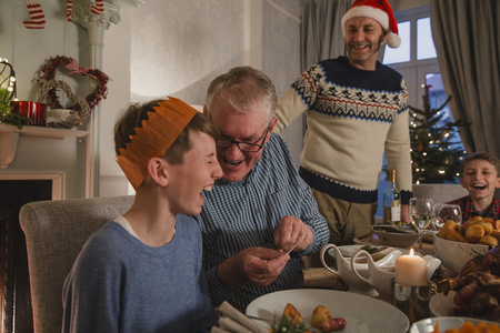 Little boy is laughing as his grandfather tells him a joke from a christmas cracker at the dinner table.の写真素材