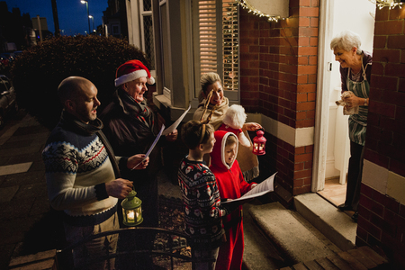 Three generation family are doing door-to-door carol singing. There is a senior woman at the door, appreciating their singing.の写真素材