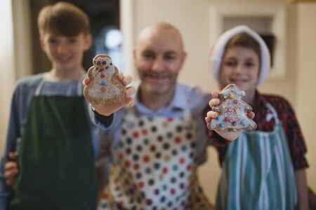 Father is standing with his two sons who are each holding up a homemade christmas biscuit to the camera.の写真素材