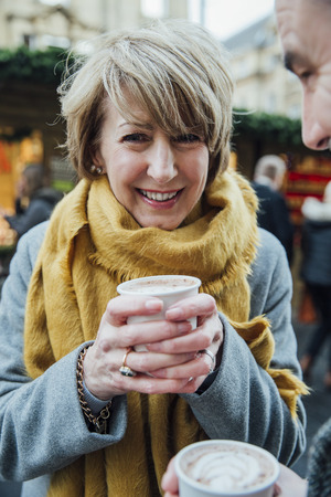 Happy mature woman is smiling for the camera while drinking a hot chocolate in the christmas market.の写真素材