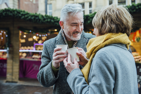 Mature couple are drinking hot drinks in a town christmas market. They are toasting the disposable cups.の写真素材
