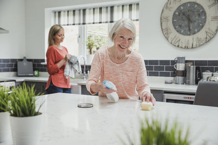 Senior woman is smiling for the camera as she cleans her kitchen with help from her daughter. の写真素材