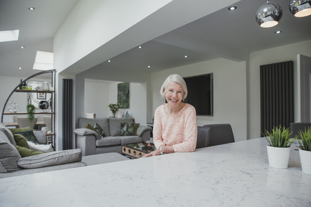 Happy senior woman is smiling for the camera while sitting at the breakfast bar in the kitchen of her home. の写真素材