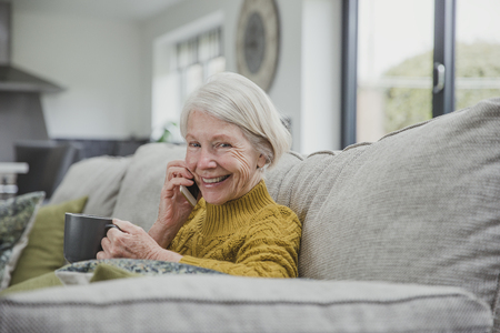 Senior woman is curled up on the sofa in her home with a cup of tea while she enjoys a phonecall on a smartphone.の写真素材