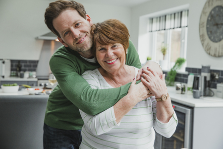 Man is posing for the camera with his mother at her home. He has his arms round her and they are both smiling for the camera.の写真素材