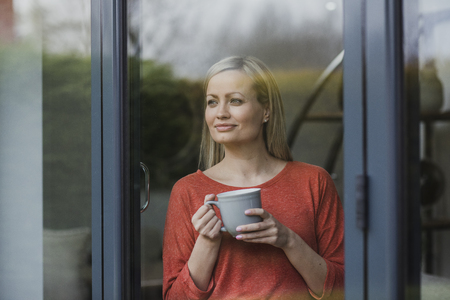 Woman is standing at the window of her home, enjoying a cup of tea.の写真素材