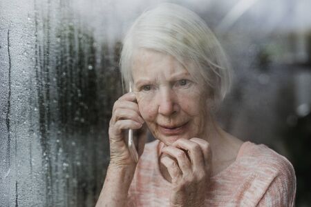 Senior woman is looking worriedly out of the window of her home while talking to someone on the phone.の写真素材