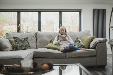 Woman is sitting on the sofa in her home, using a smart phone.の写真素材