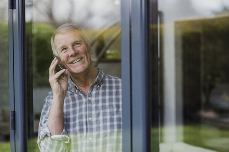 Senior man is enjoying a phone call while he looks out of a window in his home.の写真素材