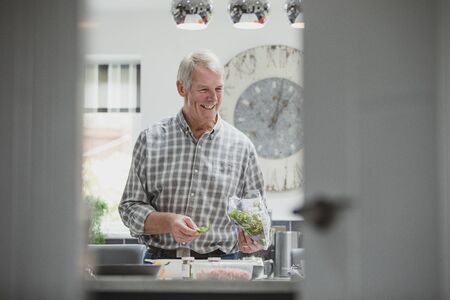 Happy senior man is preparing a meal in the ktichen of his home. He is taking rocket out of the bag and placing it in a bowl.の写真素材