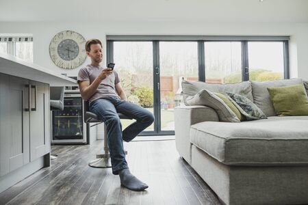 Man is relaxing in the living area of his home. He is smiling as he uses a smart phone. の写真素材