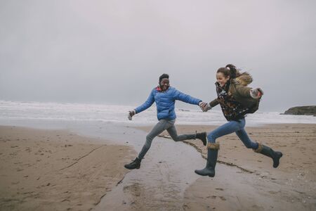 A young couple leap along a beach together in winter.の写真素材