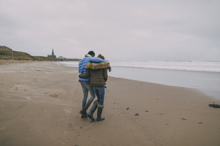 A young couple walk along a winter beach with their arms around each other.の写真素材