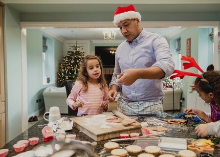 Little girls are making christmas biscuits at home with their father. Their mother is watching them form the dining table where she is working.の写真素材