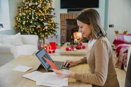 Mature woman is sitting at the dining table in her home at christmas time, writing out christmas cards. She is using her laptop for information.の写真素材