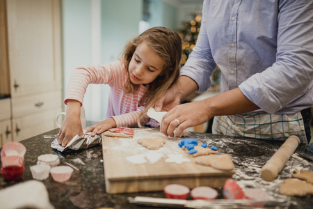 Little girl is making christmas biscuits with her dad in the kitchen of their home. They are putting icing on top of the biscuits.の写真素材