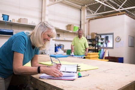 Senior businesswoman is doing paperwork in her stock warehouse. Her husband is in the background ironing fabric.の写真素材
