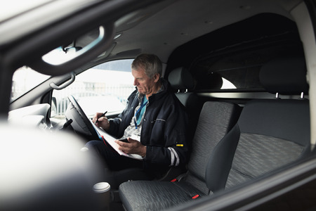 Businessman is working in the driver seat of his car while stationary. He is filling in paperwork and holding a smart phone.の写真素材