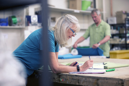 Senior businesswoman is doing paperwork in her stock warehouse. Her husband is in the background measuring fabric.の写真素材
