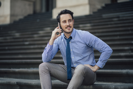 Millennial businessman is sitting on the steps of Parliament House, Melbourne, talking to someone on his smartphone. の写真素材