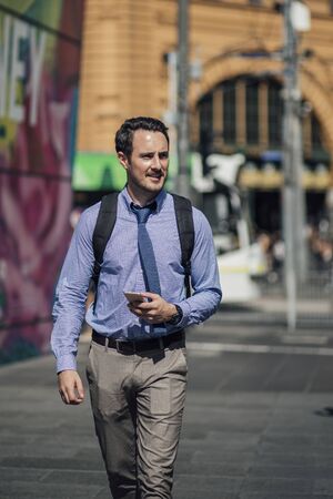 Millennial businessman is commuting to work in Melbourne, Victoria. He is walking while holding a smartphone.の写真素材