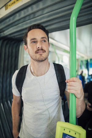 Millennial man is travelling by tram in Melbourne, Victoria. He has his headphones in and is holding a rail.の写真素材