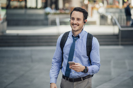 Happy millennial businessman is commuting in Melbourne, Victoria with a smartphone, listening to music through his earphones.の写真素材