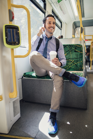 Millennial businessman is talking on the phone and holding a cup of coffee while commuting on the tram in Melbourne, Victoria. の写真素材