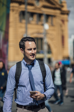 Millennial businessman is commuting to work in Melbourne, Victoria. He is walking while holding a smartphone.の写真素材