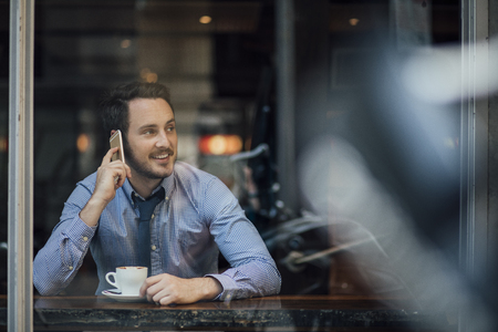 Millennial businessman is enjoying a cup of coffee in a cafe while talking to someone on his smart phone. の写真素材