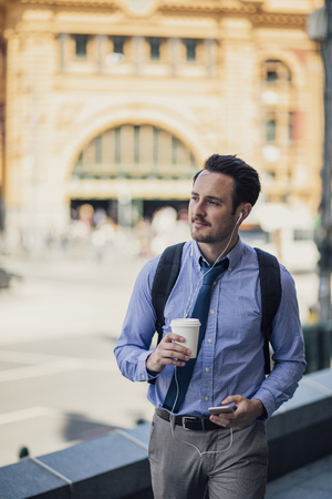 Happy millennial businessman is commuting in Melbourne, Victoria with a smartphone, listening to music through his earphones.の写真素材