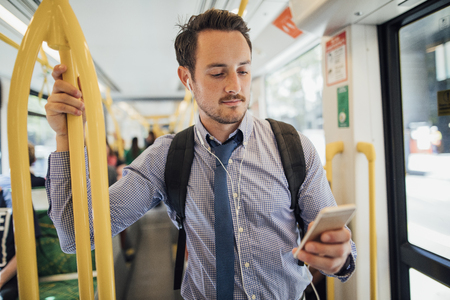 Millennial businessman is commuting on a tram in Melbourne, Victoria. He is watching something on his smart phone with headphones while standing and holding on to the rail. の写真素材