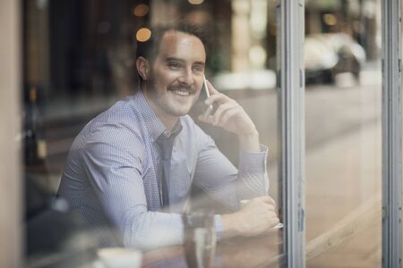 Millennial businessman is enjoying a cup of coffee in a cafe while talking to someone on his smart phone. の写真素材