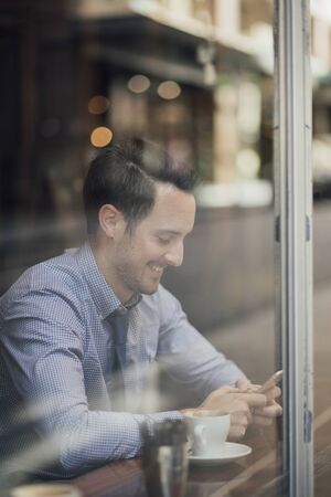 Millennial businessman is enjoying a cup of coffee in a cafe while texting on his smart phone. の写真素材