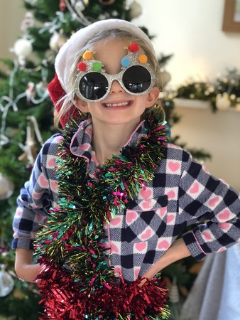 Little girl is posing for the camera in front of the christmas tree. She is wearing festive sunglasses and has tinsel wrapped round her. の写真素材