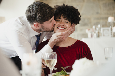 Wedding guests having fun at the meal. A woman with an afro is getting a big kiss on the cheek by the groom on his wedding day. の写真素材