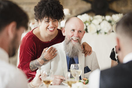 Hipster wedding guest has kissed the father of the bride with lipstick on. They are both laughing with other guests at the lipstick print she has left on his cheek. の写真素材