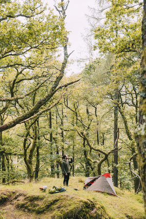 Young man is wild camping alone in the woodlands of the Lake District. He is standing outside of his tent, having a drink from a flask.の写真素材