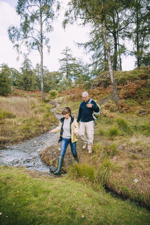 Senior couple are enjoying a walk through the woodlands together. They have come across a small stream which they are stepping over. の写真素材