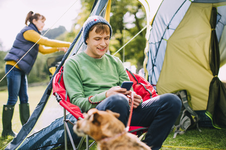 Teenage boy is sitting in a camping chair by his tent, engrossed in his smart phone while his Mum puts up the tent. の写真素材