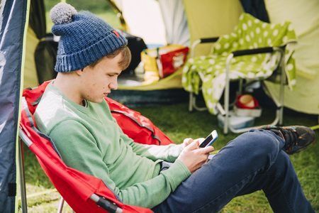 Teenage boy is sitting in a camping chair by his tent, engrossed in his smart phone. の写真素材