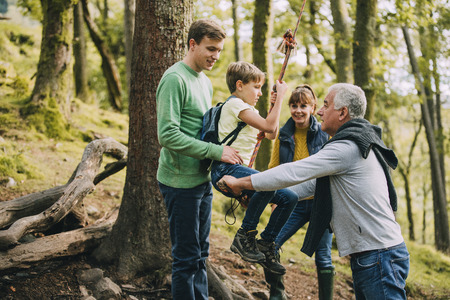 Little boy is getting on a rope swing in the forest with help from his family. They are holding him as he climbs on. の写真素材
